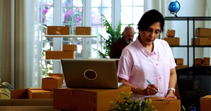 Indian senior couple doing E-commerce business from home, busy working in office managing online orders, packing parcels, dispatching boxes, taking customer calls and handling inventory together