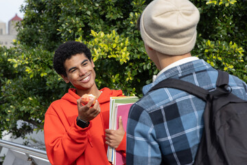 Fototapeta premium Diverse teenage male friends chatting on campus walkway, orange hoodie holding apple and notebooks