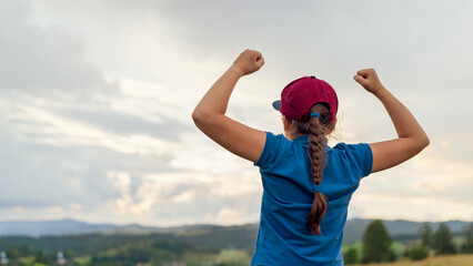 Rear view of a girl in a cap raising her arms in a gesture of victory and empowerment, standing outdoors against a scenic landscape and cloudy sky. Concept of success, confidence, and achievement.