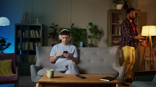 Irritated dad scolding moody teen son in headset using phone, rebellion phase