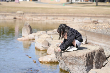 A young girl is sitting on a rock near a body of water. She is wearing a black jacket and white shoes. The scene is peaceful and serene, with the girl enjoying the calmness of the water