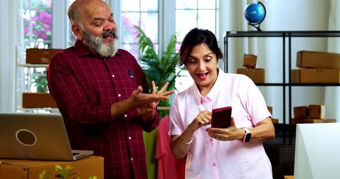 Indian senior couple celebrating success in home based E-commerce business while managing online orders, dispatching parcels, packing cardboard boxes and working together in home office