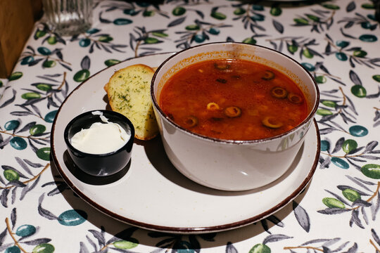Bowl of traditional Russian Solyanka soup with olives, served with garlic bread and sour cream. Rich and spicy meat soup on a floral tablecloth in a restaurant setting.