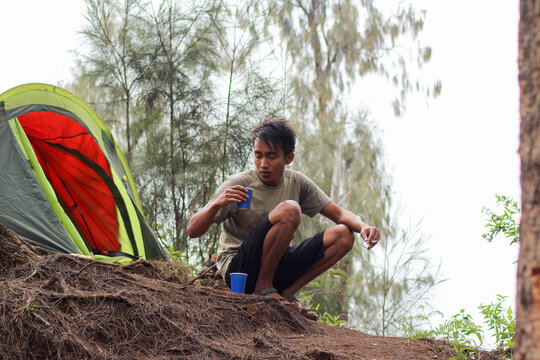 Young man enjoys drink while crouching beside tent during outdoor camping trip