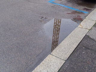 Fototapeta premium Reflection of a construction crane in a puddle on an asphalt surface with stone curb