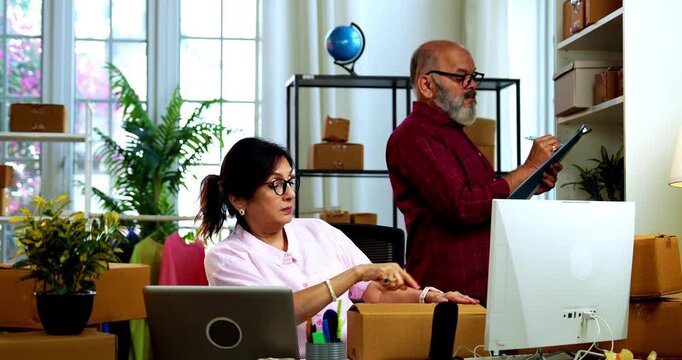 Indian senior couple packing garment while doing E-commerce business from home managing online orders, handling parcels, dispatching cardboard boxes and working happily in home office