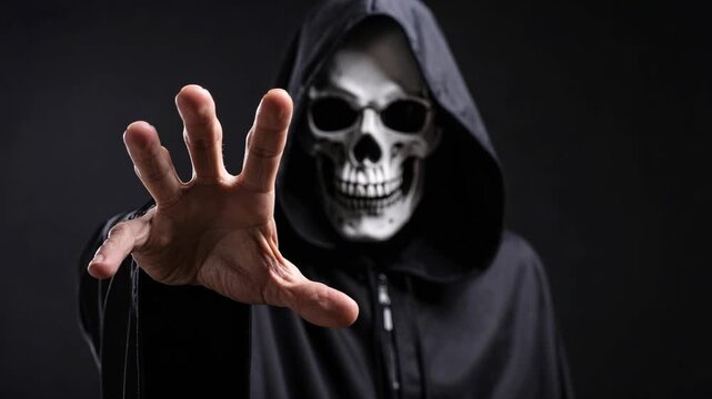 Person in a scary skull mask and black hooded cloak reaching a hand toward the camera in a dark studio