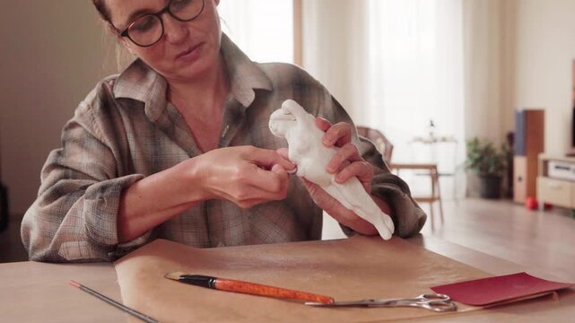 Close up of female hands polishing clay sculpture with sandpaper in 4K. Hobby sculptor sanding ceramic figurine to smooth surface in bright art studio workshop. Craft and DIY concept.
