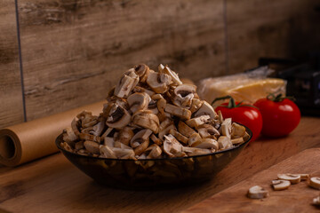 Bowl of fresh sliced mushrooms sitting on a cutting board, ready for cooking, with ripe tomatoes, a block of cheese, and baking paper in the background, showing kitchen food preparation.