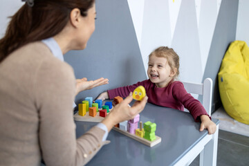 Obraz premium Preschool teacher engaging with a smiling toddler girl at a small desk, using wooden sorting blocks and a duck toy for cognitive development and speech therapy in a modern classroom