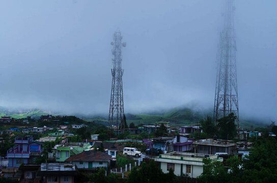 Cherrapunji, India - 27 July 2023: View of two tall cell towers rising through the mist-shrouded town, houses huddled close, green hills fading into the background.