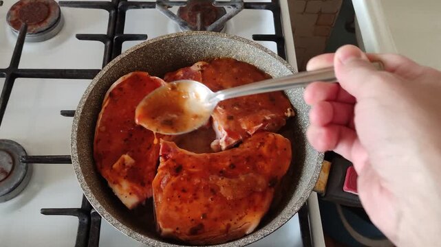 Person basting three pork pieces simmering in red sauce on frying pan with spoon, first person view video