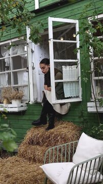A woman in black gloves and boots leans out of an open window of a green wooden house, resting her chin on her hands. Hay bales and greenery surround her, with string lights adding a cozy touch. Rusti