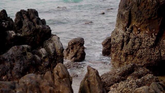Rugged rocky shoreline frames crashing waves in narrow cove with barnacle covered boulders. Calm turquoise sea contrasts jagged stone formations creating dramatic coastal texture.