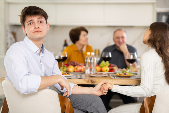 Shocked concerned young man sitting at table during family conversation with elderly parents at dinner, while girlfriend providing comfort by holding hand