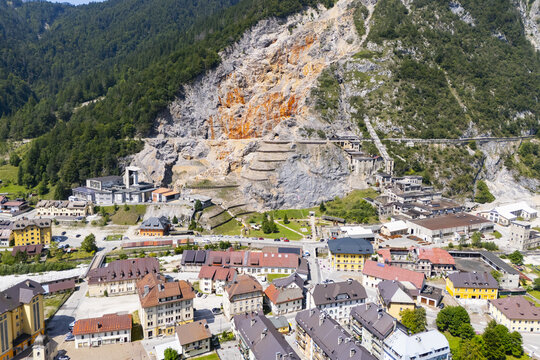 Aerial view of quaint buildings nestled against a dramatic mountain face with exposed rock and lush greenery, Cave del Predil, Friuli-Venezia Giulia, Italy.