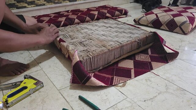 Close-up of a person upholstering a wooden seat frame with a yellow staple gun, attaching red patterned fabric during a furniture restoration project on a tiled floor