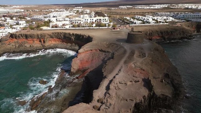 Coastal town with white buildings, rocky cliffs, a circular tower, and the ocean.