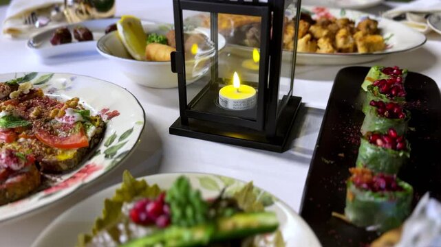 Elegant dining table setup featuring Middle Eastern appetizers, fresh salads, stuffed grape leaves with pomegranate, and a decorative candle lantern creating a warm festive ambiance indoors.