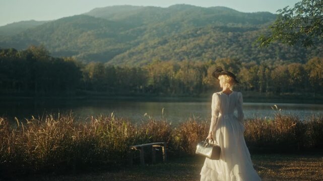Graceful Woman Dressed In White With Suitcase Taking Slow Walk Near Tranquil Mountain Shoreline