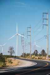 Wind turbines with high voltage electric power lines and pylons along the rural road in Thailand, carries clean alternative energy across green field landscape under blue summer sky, vertical view