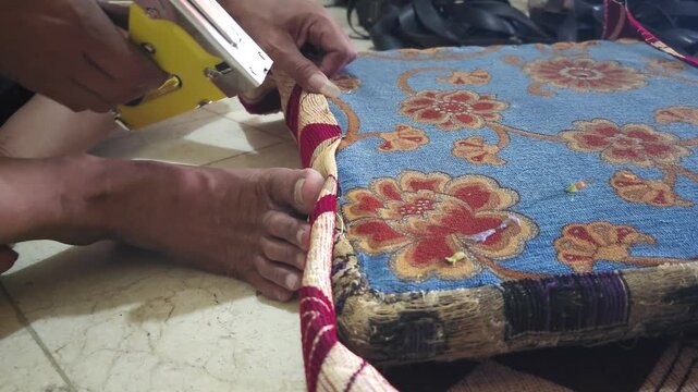 Close-up of a skilled craftsman upholstering a chair cushion with a staple gun in a workshop, securing floral fabric to the wooden frame.