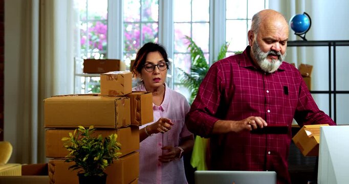 Indian senior couple making products ready to be dispatched, while doing E-commerce business from home managing online orders, packing cardboard boxes, counting garments and preparing parcels