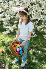 Fototapeta premium Easter egg hunt. Little girl with bunny ears holding decorative Easter egg and wicker basket under blooming spring tree. Outdoor Easter celebration. joyful childhood moment outdoors.