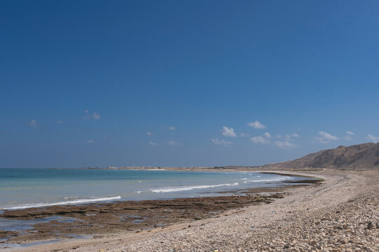 Panoramic view of a secluded Oman beach along the Mirbat-Hasik road. Clear turquoise waters meet a rocky shoreline and desert hills under a vast blue sky. Ideal for travel and nature themes.