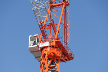 Low angle close-up bright orange tower crane cab and jib boom set against a clear blue sky.  Scene features load‑capacity sign and partial view of operator sitting in cab shaded with curtain.