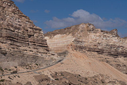 Winding asphalt road through the rugged, layered limestone cliffs of the Dhofar Mountains on the scenic drive between Mirbat and Hasik, Oman. Arid desert landscape under a clear blue sky.