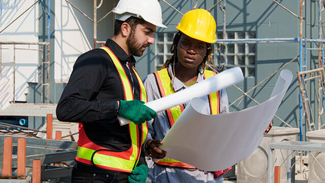 Middle Eastern male engineer and African female construction supervisor reviewing blueprint plans at outdoor building site during daytime, standing beside scaffolding structure while discussing projec