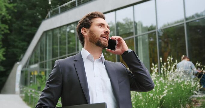 Confident businessman in a formal suit holding a laptop and talking on the phone while walking outdoors in a park area