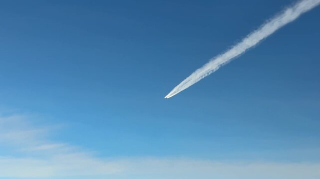 Aerial view taken from a jet airplane cockpit of another jet and its white contrail crossing in diagonal right to left the scene under a blue sky.