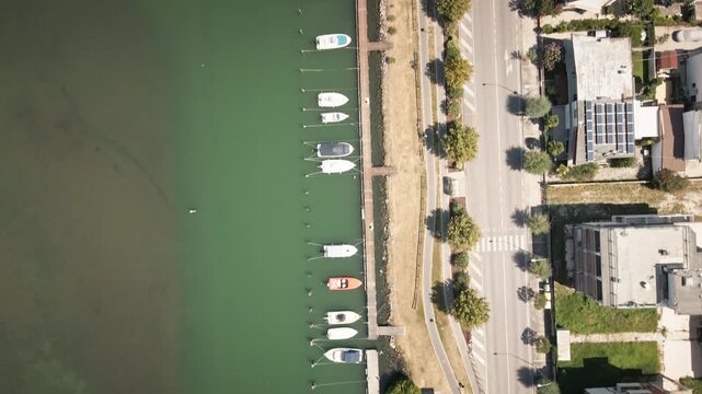 Smooth 4K UHD drone flies forward in a top-down view over Grado marina berths and waterfront road, capturing moored boats and clear lagoon water for travel ads, b-roll.