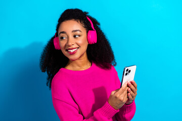 Young woman smiling wearing pink sweater and headphones holding phone against blue background...