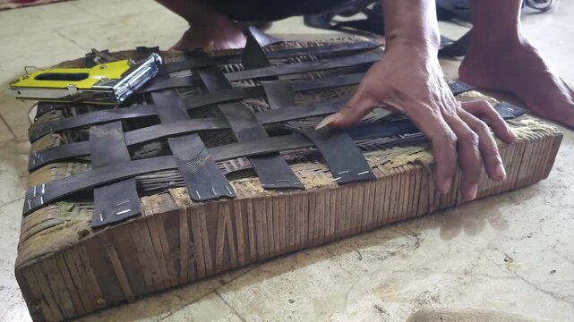 Close up of craftsman repairing wooden chair seat by weaving black rubber strips and securing with staple gun for furniture restoration