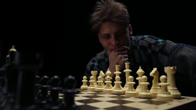 A young man is sitting in front of a chessboard with pieces and thinking about the next move. The chess player is assessing the risks before making the first move. A thoughtful chess player