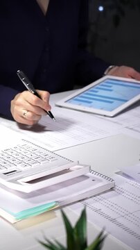 Woman working late night in office, multitasking with a tablet and writing on documents, managing heavy paperwork load. Audit and taxes in business