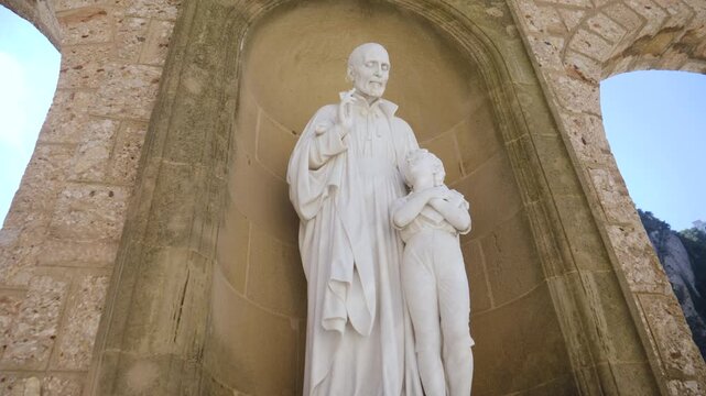 A white stone sculpture of a saint guiding a child, situated in an arched stone niche at the Montserrat monastery. This religious artwork is part of the monastery's extensive collection of outdoor sta