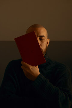A thoughtful bald man with a beard, wearing a dark turtleneck sweater, sits in moody, warm lighting with eyes closed, holding a blank red book over half his face in a conceptual, artistic pose.