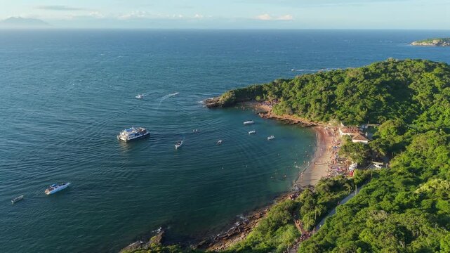 Bustling Praia da Azeda in Arma&ccedil;&atilde;o dos B&uacute;zios, Rio de Janeiro, Brazil, forming sheltered turquoise cove along South Atlantic shoreline with Mata Atl&acirc;ntica hillside and anchored boat, drone gliding