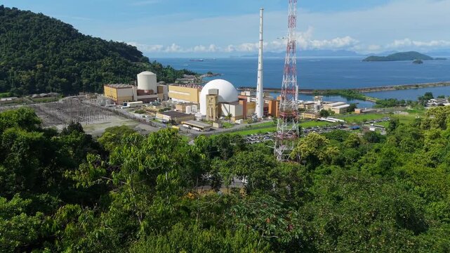 Nuclear power plant complex beside coastline with forested hills, aerial establishing overview of energy production on the coast