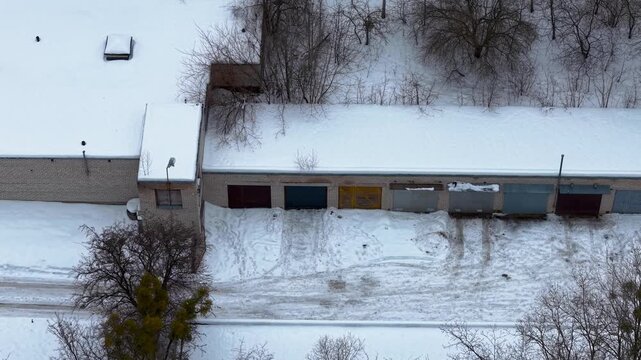 Aerial footage captures a row of snow-covered garages surrounded by bare trees in a snowy landscape. The serene winter scene, highlights the contrast between the white snow and colorful doors.
