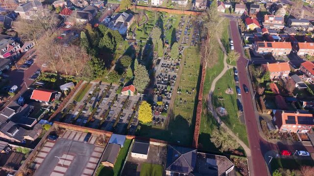 High angle aerial view of a neat European cemetery in a residential neighborhood. Features organized gravestones, lush greenery, red roofed houses, a parking lot, and a small park under soft sunlight.