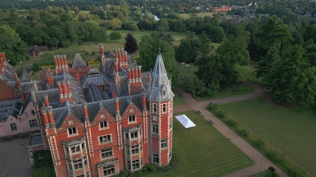An aerial view shows a grand red brick manor house with multiple chimneys and a conical tower, surrounded by lush green trees and manicured lawns.