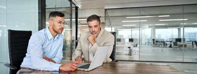 Two busy professional business men executive team, male partners working using laptop computer, manager consulting client talking analyzing online trading financial data in office sitting at desk.