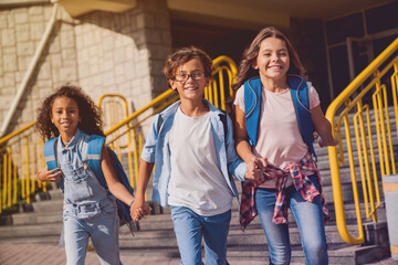 Obraz premium Happy kids elementary school pupils children with rucksacks backpack walking holding hands at school yard running out outside at the end of the lessons. Finish school and summer vacation concept.