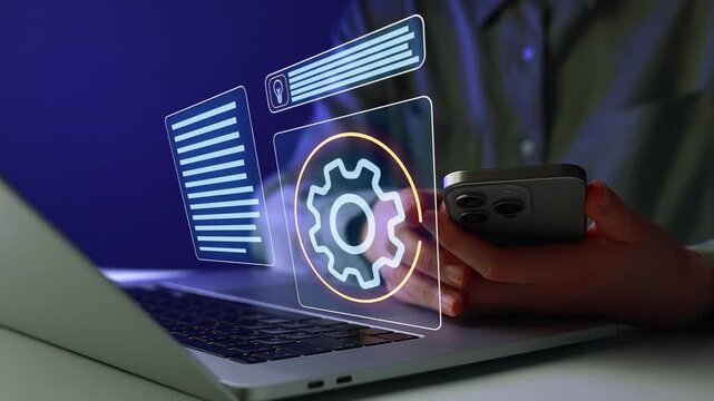 Person uses a smartphone while working on a laptop with digital elements in a dark room at night.