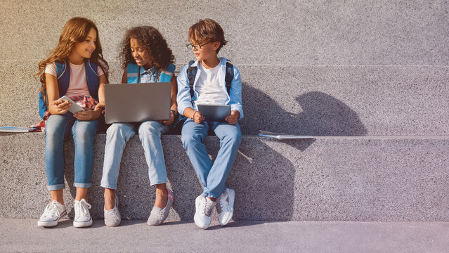 Smiling school kids preteen boys and girls elementary middle pupils children sitting on the stairs and looking in smartphone screen with digital devices laptop tablet in hands outdoors, copy space